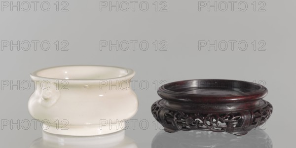 Table stand;, Shallow, recessed base on four feet. Chinese, Qing dynasty 1644–1911, 19th century. Geometric and plant ornament. Height 3.2 cm; diameter 9.4 cm., A glossy off-white ceramic vessel with a rounded body, a slightly flared rim, and a small raised decorative knob on its side sits next to a low, dark wooden stand with a circular top and an intricately carved openwork skirt; the ceramic piece is pale cream with subtle sheen and tiny darker specks, while the wooden stand is deep brown to reddish-brown with a polished finish, both objects resting on a reflective surface against a smooth light gray background.