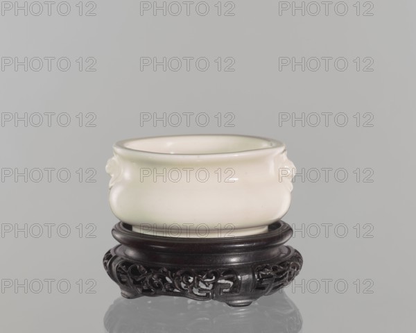 Table stand;, Chinese; Qing dynasty 1644–1911; 19th century. Shallow, recessed base on four feet. Geometric and plant ornament. Height 3.2 cm; diameter 9.4 cm., A smooth rounded ceramic bowl with a glossy off-white surface sits on an ornate dark wooden stand; the bowl has subtle rounded protrusions on its sides and a slightly flared rim, and the stand features carved openwork patterns and a dark brown to nearly black finish against a plain light gray background with a faint reflection below.
