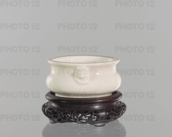 Table stand;, Shallow, recessed base on four feet. 19th century, Qing 1644–1911, China. Material/decoration: geometric and plant ornament. Height 3.2 cm; diameter 9.4 cm., A small rounded ceramic bowl in a creamy white color sits on a dark wooden stand with carved openwork. The bowl has a slightly flared rim and a subtle raised decorative motif on its side in the same creamy white color. The wooden stand is a deep brown to almost black color with smooth circular tiers above an intricately carved base. The background is a uniform light gray, and a soft reflection of the stand and bowl appears on the glossy surface beneath them.