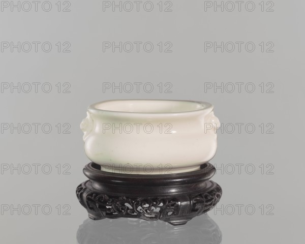 Table stand; unknown creator; gift; Chinese art; Chinese; Qing dynasty 1644–1911; geometric and plant ornament; shallow, recessed base on four feet; 19th century; China; height 3.2 cm, diameter 9.4 cm., A small rounded bowl with a smooth glossy surface in off-white and pale cream tones sits on a dark carved wooden stand; the bowl has subtle curved handles on its sides and a slightly flared rim, and the stand is deep brown to nearly black with ornate openwork carving and a circular top, all shown against a plain pale gray background.