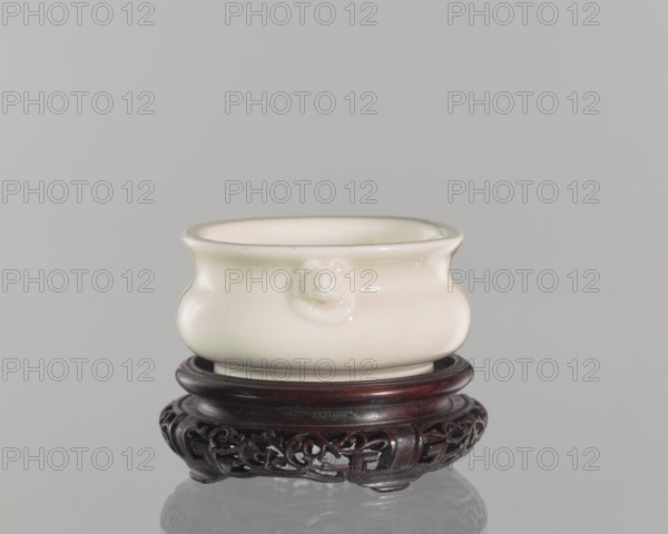 Table stand, shallow recessed base on four feet; geometric and plant ornament; Chinese; Qing dynasty 1644–1911; 19th century; height 3.2 cm, diameter 9.4 cm., A small rounded ceramic vessel sits on an ornate carved wooden stand; the vessel is a glossy off-white or cream color with a subtle sheen and a slightly flared rim, and it has a raised decorative feature on its side. The stand is a dark reddish-brown wood with intricate openwork carving and a polished finish. The background is a smooth light gray, and a faint reflection of the stand and vessel appears on the surface beneath them.