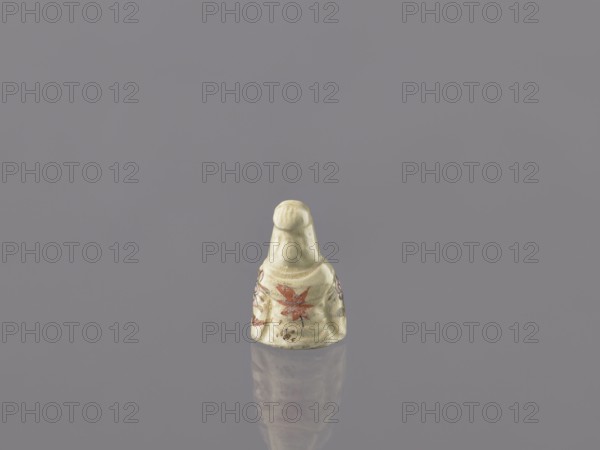 Seated female figure — Guanyin, gift; early 20th century, Japan; ceramic sculpture; height 3.7 cm, width 2.4 cm; depicts a female image/figurine, Japanese art., A small ceramic figurine sits centered on a smooth reflective surface against a gray background, showing an off-white or cream color overall with painted accents in red and muted green; the figurine has a rounded top and a flared base with floral-like motifs on its sides, and its reflection is visible beneath it on the surface.