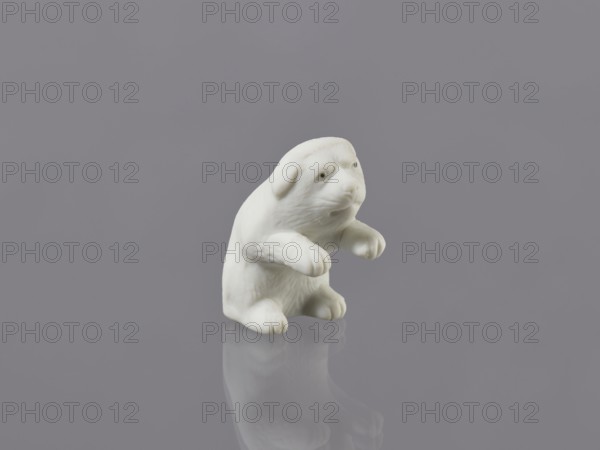 Puppy, unknown ceramicist, Meiji period early 20th century, Meiji 1868–1912, Japan — Hirado. Porcelain figurine of a dog. Height 7.0 cm; width 3.6 cm; weight 46.0 g., A small white sculpted figure of an animal with rounded ears and a snout sits on a smooth reflective surface, showing a soft reflection beneath; the figure has rounded paws held slightly forward and textured fur details. The background and surface are shades of gray and muted lavender, and the overall palette includes white and several tones of gray.