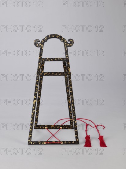 Mirror stand kagami-kake, Japanese, Edo period 1603–1868, lacquer, wooden stand covered with black lacquer; painted with gold with plant ornaments, 1st half of the 19th century, Japan, stand, height 66 cm, A decorative black wooden frame with a tapered triangular shape stands against a light gray background, decorated with a repeating gold floral and vine pattern; the frame has a curved top with scroll-like ends and a few horizontal crossbars, and a red cord threaded near the base forming a crossed pattern while several red tasseled cords lie beside the frame.
