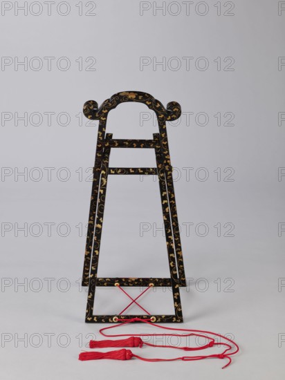 Mirror stand kagami-kake, wooden stand covered with black lacquer; painted with gold with plant ornaments, mirrors, Edo period 1st half of the 19th century, Japan, height 66 cm, A decorative black wooden stand with curved top elements and scroll-like ends, covered in a gold floral and vine pattern, standing on a light gray surface against a pale gray background, with a few red cords and tassels resting in front of it.