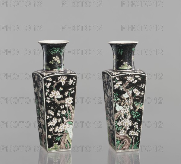 Pair of vases, Chinese, 18th century, colored porcelain famille noire, decorated with birds, plums, and floral motifs; vessel, height 22.5 cm., A pair of tall, square-based vases with flared necks stand side by side against a plain gray background, each decorated with a predominantly black surface covered in floral and tree branch motifs in white, green, and brown tones; the flared rims and neck interiors are white, and the painted scenes include pale green leaves and accents along with brown tree trunks and white blossoms, with reflections visible beneath the vases on the glossy surface.