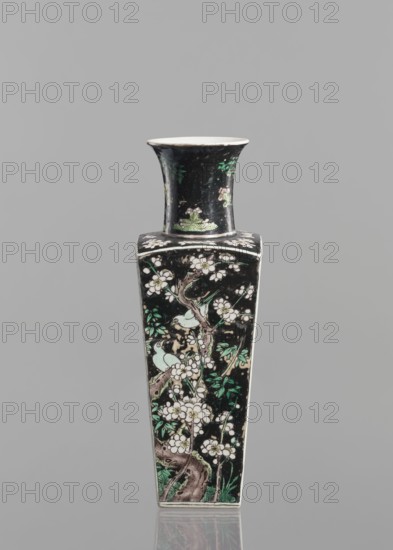 Pair of vases, Chinese, 18th century, colored porcelain famille noire, decorated with birds, plums and floral motifs; vessel, height 22.5 cm., A tall, narrow ceramic vase with a flared rim and a square shoulder, decorated on a dark background with floral and bird motifs; the surface shows branches with blossoms in white and pale pink, leaves in shades of green, and pale green birds perched among the flowers, with brownish tree trunks and subtle green foliage at the base; the rim and interior appear white, and the overall background is black with some lighter worn areas, set against a neutral gray backdrop.