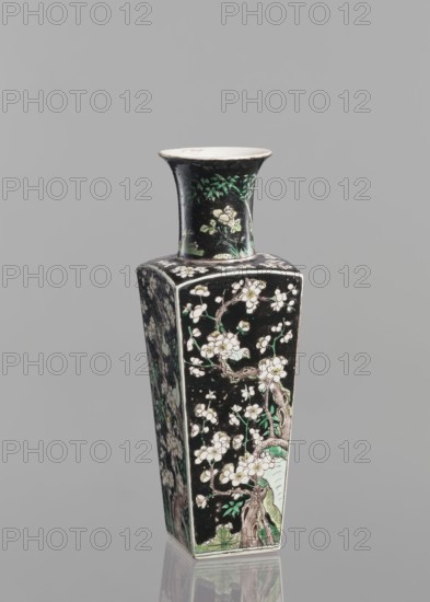 Pair of vases; Chinese; 18th century; colored porcelain famille noire; decorated with birds, plums, and floral motifs; vessel; height 22.5 cm; ., A tall, square-based ceramic vase with a flared rim, shown against a plain gray background. The vase has a predominantly black surface decorated with floral and tree branch motifs in white, green, and pale pink, with brownish outlines and some lighter green foliage near the base and neck. The neck features a cluster of green leaves and pale flowers, while the body panels display blossoming branches and leaves in white, green, and pale pink set against the black ground. The rim interior and the very top edge are a pale off-white color. The vase sits on a reflective surface that shows a faint mirror image of its lower section.