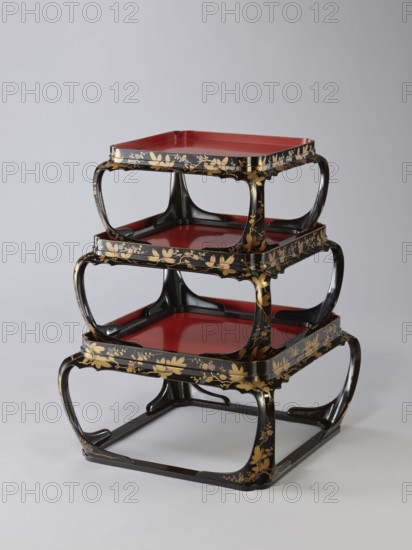 Set of three trays on legs kakeban. Japanese, Edo period early 19th century. Low tables, identically decorated but of different dimensions allowing them to be stacked pyramidally, one on top of the other. Each table has an almost square top framed with high slats, supported by four curved legs connected at the bottom by four slats forming a square base. The top and the inner side of the slats framing it are coated with vermilion-coloured lacquer; the remaining surfaces are covered with shiny black roiro-nuri lacquer and decorated with a golden ornament of pine branches and plum blossoms made using the sprinkling technique hiramaki-e, the decoration slightly raised above the background surface. Subject motifs: plum trees, flowers, trees, pine trees, twigs, winter. Piece of furniture., A stacked set of three square trays with rounded openwork frames, each tray having a glossy red interior and a black exterior decorated with gold floral motifs; the frames and legs are black with flowing c...