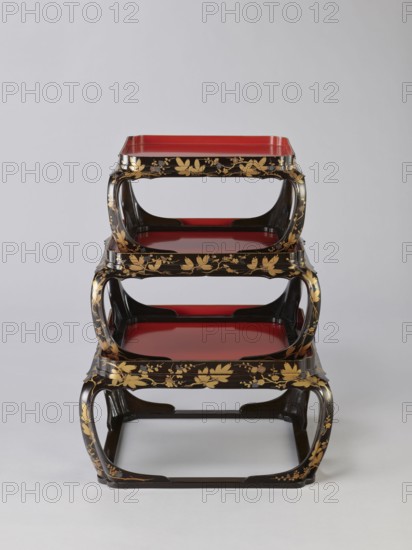 Set of three trays on legs kakeban, maker, early 19th century Edo period, Japan. Low tables, identically decorated but of different dimensions allowing them to be stacked pyramidally, one on top of the other. Each table has an almost square top framed with high slats, supported by four curved legs connected at the bottom by four slats forming a square base. The top and the inner side of the slats framing it are coated with vermilion-coloured lacquer; the remaining surface is covered with shiny black roiro-nuri lacquer and decorated with a gold ornament of pine branches and plum blossoms made using the sprinkling technique hiramaki-e, the decoration slightly raised above the background surface. Subject motifs: plum trees, flowers, trees, pine trees, twigs, winter. Piece of furniture., A stack of three nested rectangular trays with rounded open sides, each tray showing a glossy red surface framed by black edges decorated with gold floral and leaf motifs, the frames forming curved support...