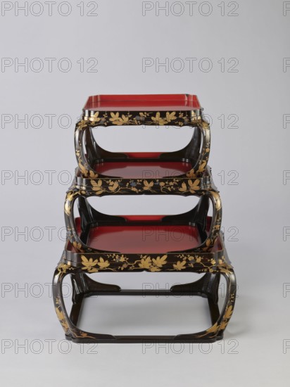 Set of three trays on legs kakeban, Japanese, Edo period early 19th century. Low tables, identically decorated but of different dimensions allowing them to be stacked pyramidally, one on top of the other. Each table has an almost square top framed with high slats, supported by four curved legs connected at the bottom by four slats forming a square base. The top and the inner side of the slats framing it are coated with vermilion-coloured lacquer; the remaining surface is covered with shiny black roiro-nuri lacquer and decorated with a golden ornament of pine branches and plum blossoms made using the sprinkling technique hiramaki-e, the decoration slightly raised above the background surface. Piece of furniture., A stacked set of three lacquered trays with open curved frames, each tray showing a red flat surface bordered by black edges; the frames are black with gold floral and leaf decoration, and the overall piece sits against a light gray background with visible shadows.