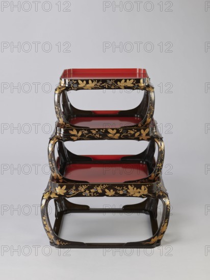 Set of three trays on legs kakeban. Japanese, Edo period early 19th century. Low tables, identically decorated but of different dimensions allowing them to be stacked pyramidally, one on top of the other. Each table has an almost square top framed with high slats, supported by four curved legs connected at the bottom by four slats forming a square base. The top and the inner side of the slats framing it are coated with vermilion-coloured lacquer; the remaining surfaces are covered with shiny black roiro-nuri lacquer and decorated with a golden ornament of pine branches and plum blossoms made using the sprinkling technique hiramaki-e; decoration slightly raised above the background surface. Subject motifs: plum trees, flowers, trees, pine trees, twigs, winter. Piece of furniture., A three-tiered stand with curved rectangular frames appears centered against a pale gray background, the stand showing a glossy black finish with decorative gold floral and leaf patterns along the edges and su...