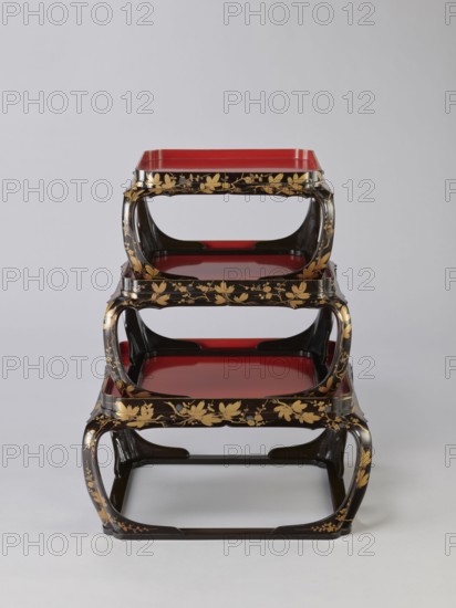 Set of three trays on legs kakeban, Japanese, Edo period early 19th century. Low tables, identically decorated but of different dimensions allowing them to be stacked pyramidally, one on top of the other. Each table has an almost square top framed with high slats, supported by four curved legs connected at the bottom by four slats forming a square base. The top and the inner side of the slats framing it are coated with vermilion-coloured lacquer; the remaining surface is covered with shiny black roiro-nuri lacquer and decorated with a golden ornament of pine branches and plum blossoms made using the sprinkling technique hiramaki-e; decoration slightly raised. Subjects: plum trees, flowers, trees, pine trees, twigs, winter. Function: piece of furniture., A stack of three nested trays with curved open sides sits against a plain light gray background; each tray has a glossy red interior and a glossy black exterior decorated with gold floral and leaf motifs, with the black frames forming r...