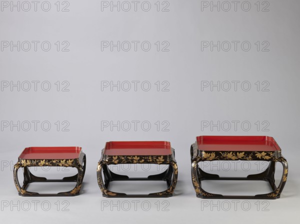 Set of three trays on legs kakeban, Japanese, Edo period early 19th century. Low tables, identically decorated but of different dimensions allowing them to be stacked pyramidally, one on top of the other. Each table has an almost square top framed with high slats, supported by four curved legs connected at the bottom by four slats forming a square base. The top and the inner side of the slats framing it are coated with vermilion-coloured lacquer; the remaining surface is covered with shiny black roiro-nuri lacquer and decorated with a golden ornament of pine branches and plum blossoms, made using the sprinkling technique hiramaki-e; decoration slightly raised above the background surface. Subject: plum trees, flowers, trees, pine trees, twigs, winter. Piece of furniture., A row of three low square lacquered stands arranged from left to right, each with a red flat top surface and a glossy black base frame; the black frames feature gold painted floral and leaf motifs along their sides an...