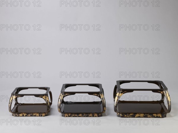 Set of three trays on legs kakeban. Japanese, Edo period, early 19th century. Low tables, identically decorated but of different dimensions allowing them to be stacked pyramidally. Each has an almost square top framed with high slats, supported by four curved legs connected at the bottom by four slats forming a square base. The top and the inner side of the slats framing it are coated with vermilion-coloured lacquer; the remaining surface is covered with shiny black roiro-nuri lacquer and decorated with a golden ornament of pine branches and plum blossoms made using the sprinkling technique hiramaki-e; decoration slightly raised above the background surface. Subject: plum trees, flowers, trees, pine trees, twigs, winter. Piece of furniture., Three low, rectangular stands sit side by side against a neutral light gray background, each with a glossy dark brown or black lacquer finish and curved open sides. The stands form a tiered shape with a flat top frame and a lower shelf, and they fe...