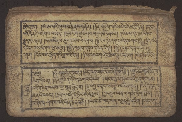 Buddhist text - Commentary on the praise of the names of Manjushri by Ga Rabjampa Kunga Yeshe Tib. 'phags pa 'jam dpal gyi mtshan yang dag par brjod pa zhugs so, unknown creator role, author. Buddhist art, Tibetan, graphic prints, Book in Tibetan pecha style, xylographic print in student script dbu can. 26 folios, recto page numbering, first verso, 2 pages missing. Buddhist commentary on the prayer of praise in honor of the names of the bodhisattva Manjushri Tib. 'jam dpal dbyang by Ga Rabjampa Kunga Yeshe Tib. sga rab 'byams pa kun dga' ye shes. Original text from the 15th century; object dated 19th century, Tibet. Graphics, width 24.6 cm, height 8.0 cm, A worn rectangular manuscript page lies flat with a horizontal fold across the middle; the page surface is textured and aged, showing tones of beige, tan, and brown with darker brown edges and some darker smudging. Two bordered rectangular blocks occupy the upper and lower halves, each filled with dense dark markings arranged in rows;...