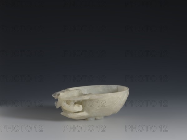 A vessel for rinsing brushes, Chinese, Qing dynasty 1644–1911, 19th century. Materials/subject: painting implements; peaches iconography. Function: vessel. Dimensions: height 3.5 cm, width 11.0 cm, depth 7.4 cm., A pale, translucent bowl with smooth, slightly irregular surfaces rests on a flat surface, showing a sculpted handle-like extension with flowing, interlocking forms on one side; the bowl's material appears milky white with subtle variations in translucency and texture, and it casts a soft shadow on the light-colored foreground while a dark gradient background transitions from deep charcoal at the top to a softer gray near the horizon.