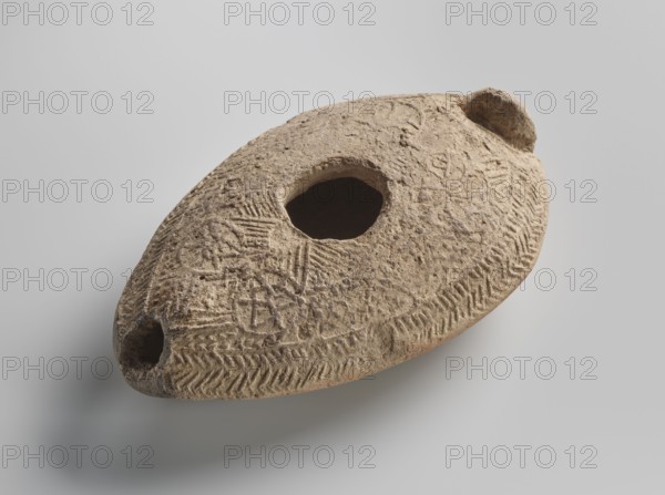 Oil lamp, Islamic art, Arabic culture, Umayyad dynasty 661–750, Hisham II Umayyad caliph; 691–743, utility ceramics, Arabic ceramics, oil lamp, everyday life in the Middle East, geometric ornament, made of fine yellowish clay, modelled in a two-part mould; the upper part, with relief patterns, is decorated with rosette motifs arranged symmetrically among delicate dots; the entire composition at the edge of the lamp is surrounded by an ornamental band filled with a chevron pattern. 7th–8th century, Khirbat al-Mafjar, height 3.2 cm, width 6.9 cm, length 11.0 cm, A roughly oval, beige-brown clay object with a textured, pitted surface sits against a light gray background; it has a large irregular central opening and a smaller rounded opening near one tapered end, with shallow incised linear and chevron patterns across its top and edges, and an overall weathered, earthy appearance with variations of tan, light brown, and darker brown in the crevices.