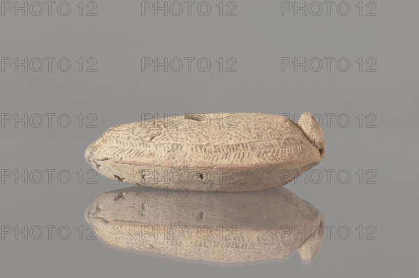Oil lamp, Islamic art, Arabic culture, Umayyad dynasty 661–750, Hisham II Umayyad caliph; 691–743, utility ceramics, Arabic ceramics, oil lamp used in everyday life in the Middle East, geometric ornament. Made of fine yellowish clay, modelled in a two-part mould; the upper part, with relief patterns, is decorated with rosette motifs arranged symmetrically among delicate dots. The entire composition at the edge of the lamp is surrounded by an ornamental band filled with a chevron pattern. 7th–8th century, found at Khirbat al-Mafjar. Height 3.2 cm, width 6.9 cm, length 11.0 cm., A small, rounded object with a flattened oval shape rests on a reflective surface, showing a mirrored image below; the object is textured with carved, repeating patterns and shallow grooves across its top and sides, and it has an irregular, slightly raised rim and a small protrusion at one end; the surface appears rough and weathered with tiny pits and cracks. The visible colors include beige, tan, light brown, a...