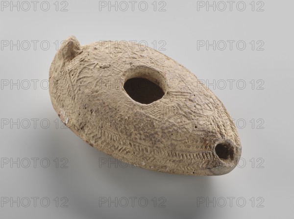Oil lamp, Islamic art, Arabic culture, Umayyad dynasty 661–750, Hisham II Umayyad caliph; 691–743, utility ceramics, Arabic ceramics, oil lamp used in everyday life in the Middle East, geometric ornament. Made of fine yellowish clay, modelled in a two-part mould; the upper part was decorated in relief with rosette motifs arranged symmetrically among delicate dots. The entire composition at the edge of the lamp is surrounded by an ornamental band filled with a chevron pattern. 7th–8th century, found at Khirbat al-Mafjar. Height 3.2 cm, width 6.9 cm, length 11.0 cm., An oval, disc-like object with a rough, textured surface sits against a pale gray background; it is primarily beige with variations of light brown and sandy tones, showing darker brown areas around some edges and recesses. The object has a large central circular opening and a smaller circular opening near one tapered end, and a raised knob-like projection near the opposite side. The surface is marked with shallow incised pat...