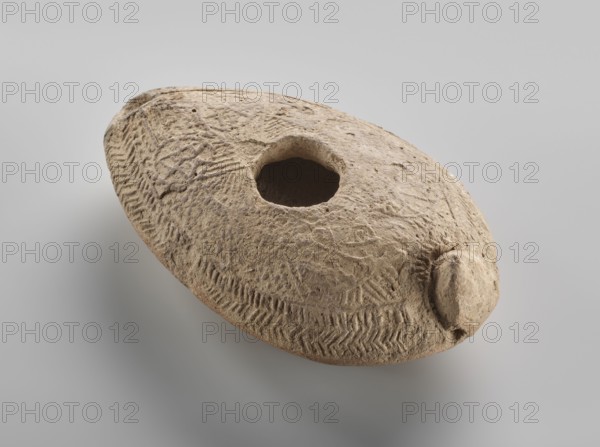 Oil lamp, Islamic art, Arabic culture, Umayyad dynasty 661–750, Hisham II Umayyad caliph; 691–743, utility ceramics, Arabic ceramics, oil lamp used in everyday life in the Middle East, geometric ornament. Made of fine yellowish clay, modeled in a two-part mould; the upper part is decorated in relief with rosette motifs arranged symmetrically among delicate dots. The entire composition at the edge of the lamp is surrounded by an ornamental band filled with a chevron pattern. 7th–8th century, found at Khirbat al-Mafjar. Height 3.2 cm, width 6.9 cm, length 11.0 cm., A round, flattened stone object with a central circular hole and a small protrusion on one side, showing a rough, textured surface with carved patterns and incised markings; the object appears in shades of beige and light brown against a smooth gray background.