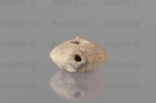 Oil lamp, Islamic art, Arabic culture, Umayyad dynasty 661–750, Hisham II Umayyad caliph; 691–743, utility ceramics, Arabic ceramics, oil lamp used in everyday life in the Middle East, geometric ornament, made of fine yellowish clay, modelled in a two-part mould; the upper part is decorated in relief with rosette motifs arranged symmetrically among delicate dots, the composition at the rim is surrounded by an ornamental band filled with a chevron pattern. 7th–8th century, found at Khirbat al-Mafjar; height 3.2 cm, width 6.9 cm, length 11.0 cm, A small, rounded, oval-shaped object with a textured, rough surface sits centered against a smooth gray background and is reflected faintly below; the object appears in shades of beige, tan, light brown, and pale gray, with a darker circular opening visible on one side and a small raised knob on the top, and the surface shows carved or incised markings and irregularities in color and texture.