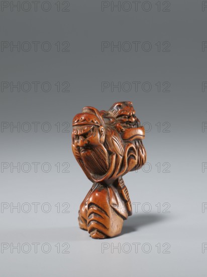 Netsuke in the shape of Shôki and an oni, unknown sculptor; Japanese, Edo period 1603–1868, 1st half of the 19th century. Carved netsuke depicting Shôki mythology and a demon oni; materials: sculpture/netsuke; height 4.5 cm, width 3.0 cm., A small carved object in warm brown tones stands on a smooth light gray surface against a darker gray background; the carving depicts a stylized figure with a detailed face, long carved beard, and a patterned headband, and there is a second carved face or figure positioned behind and slightly above the first; the surface shows carved grooves and textured lines creating flowing patterns on the body and base, with highlights and shadows emphasizing the polished, slightly glossy finish and darker recessed areas.