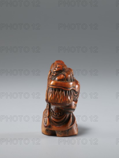 Netsuke in the shape of Shôki and an oni, Japanese, Edo period 1603–1868, 1st half of the 19th century. Carved netsuke figurine depicting the demon queller Shôki and an oni; materials: sculpture netsuke. Dimensions: height 4.5 cm, width 3.0 cm., A small carved figure in shades of brown with darker brown accents sits on a smooth light gray surface against a gradient gray background, showing carved hair and draped clothing details and a rounded top, with glossy and matte wooden textures visible.
