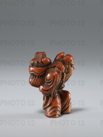 Netsuke in the shape of Shôki and an oni, Japanese, Edo period 1st half of the 19th century, netsuke sculpture, ivory or unspecified material, height 4.5 cm, width 3.0 cm; subject: Shôki mythological figure and demon/oni; function: netsuke/figurine., A small carved wooden figure in reddish-brown tones stands against a smooth gray background; the figure shows a bearded person wearing draped robes with flowing carved folds and carrying a textured basket or bundle on their back, with carved hair and facial features visible and darker carved recesses accenting contours and creases, and a soft shadow beneath the figure on the surface.