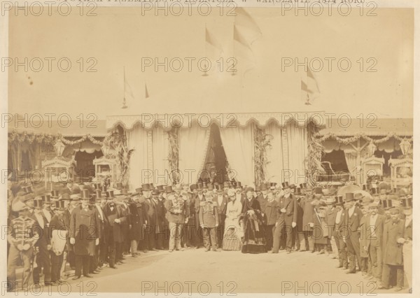 Industrial and Agricultural Exhibition at Ujazdowski Square in Warsaw in 1874. A group of people in front of the Count's pavilion. Ludwik Krasinski during the opening on September 15. Fajans, Maksymilian 1825–1890 author/photographer. Exhibitions; Industrial and Agricultural Exhibition Warsaw; 1874; exhibition pavilions; Warsaw Masovian Voivodeship; Ujazdów Warsaw; industrial and economic exhibitions; Krasinski, Ludwik 1833–1895; Kotzebue, Pawel 1801–1884; Krasinski, Ludwik 1833–1895 — iconography; Kotzebue, Pawel 1801–1884 — iconography. Print glued to cardboard with the entire surface. 15/09/1874, Warsaw. Photograph, photograph from nature. Photographic print: height 26.9 cm, width 17.9 cm; cardboard: height 30.5 cm, width 20.7 cm; passe-partout: height 43.0 cm, width 32.0 cm., A large group of people stand in front of a decorated tent with scalloped trim and draped curtains, forming a loose semi-circle. Many of the people wear dark suits and top hats, and a few wear lighter clothing...