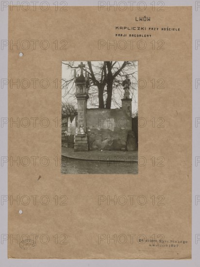 Chapel at the Church of St. Mary Magdalene in Lviv, with sculpture of John of Nepomuk on the wall of the former Dominican church; photograph by Józef Kosciesza Jaworski fl. ca. 1911–ca. 1929; negative dated 1911–1918, print dated 1911/1918–ca. 1922; photographic print glued to brown cardboard print: 15.7 × 10.6 cm; support: 35.2 × 25.6 cm; photography, nature photography., A sepia-toned photograph mounted on a brown paper sheet shows an outdoor scene with a weathered wall and a gate structure; the central image features a stone pillar topped with a cross on one side and a statue on a pedestal on the other, with bare tree branches rising behind them. The ground in front appears to be a paved street or sidewalk with a curved curb, and the wall surface shows patches of peeling plaster and uneven texture. A few posters or paper remnants are visible on a section near the left pillar. The sheet around the photograph is a warm brown color with a slightly rough texture and small punched holes ...