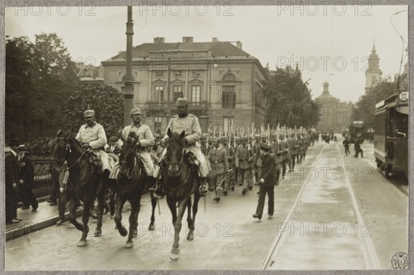 1. Warsaw. German cavalry in the street, surrounded by children, August 5, 1915; 2. Warsaw. German regiment and people on Krakowskie Przedmiescie, near Zamkowy Square, August 5, 1915; 3. German troops marching along Krakowskie Przedmiescie, past the Tyszkiewicz-Potocki Palace. Photographer: Alfred Kühlewindt 1870–1945. Three photographs glued together on a single cardboard; 5 August 1915. Subjects and details: occupation, World War I 1914–1918, Warsaw Masovian Voivodeship, trams, Krakowskie Przedmiescie street, German army, horses, palaces, soldiers, children, Warsaw inhabitants, Sigismund's Column, Staszic Palace, Church of the Holy Cross, St. John the Baptist. Photography; Poland; Warsaw., A sepia-toned street scene shows a mounted group of men in uniform riding horses at the front of a marching formation, with a larger group of uniformed soldiers walking behind them holding long rifles. A few civilians stand or walk alongside the street, and a tram or streetcar is visible on the rig...