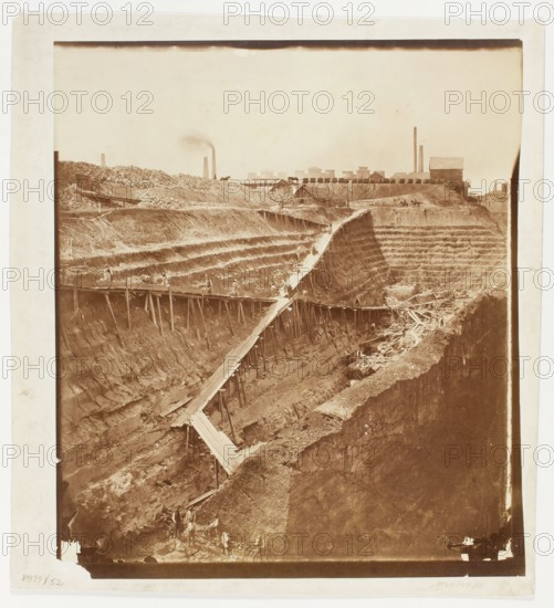 Open-pit hard coal mine Cieszkowski in Dabrowa Górnicza - view from the east, Fajans, Maksymilian 1825-1890 photographer, Light-sensitive, coal mines, Art of photography, Dabrowa Górnicza Silesian Voivodeship, miners, industrial landscapes, Zaglebie Dabrowskie Silesian Voivodeship, Print glued to cardboard with its entire surface, 1864 references to the taking of the photograph: Tygodnik Ilustrowany, 1864, no. 256 of August 20, p. 307; Gazeta Warszawska, 1864, no. 192 of August 23, p. 1, Dabrowa Górnicza place of origin, nature photography, photograph, photographic print: height 42.5 cm, width 37.7 cm; cardboard: height 47.2 cm, width 42.9 cm, A sepia-toned photograph showing a large excavated pit with stepped, terraced walls and exposed soil layers, a long wooden plank ramp supported by vertical posts zigzagging down into the pit, a few small figures of people working along the ramp and at the bottom, scattered wooden beams and debris in the lower area, a raised flat area at the top w...