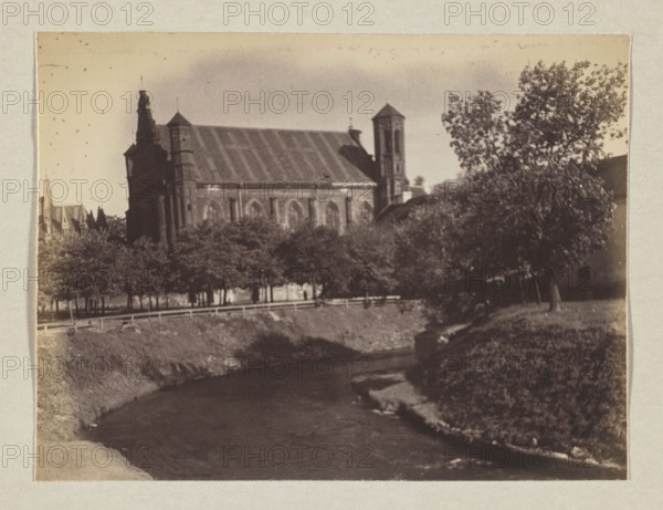 Bernardine Church; photograph, 1912; photograph, photographic print: height 11.3 cm, width 15.1 cm; album page: height 31.0 cm, width 23.5 cm; maker: Jan Bulhak 1876–1950; place: Vilnius Lithuania., A sepia-toned photograph shows a large stone building with pitched roofs and a tower partially obscured by a group of trees in front; the building appears darker brown against a lighter brown sky. In the foreground a curving river with darker brown water flows between grassy banks that are medium brown and lighter brown in places. A row of trees with greenish-brown foliage lines the far bank near a pale brown railing, and a taller tree with darker green-brown leaves stands to the right. The overall palette includes shades of brown, beige, and greenish-brown.