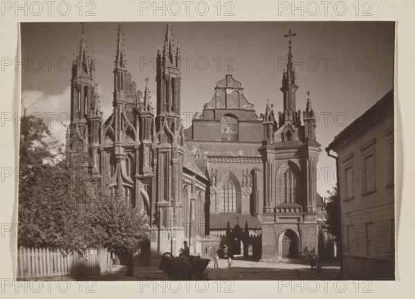 Bernardine Church; photograph, 1912; photographer Jan Bulhak 1876–1950; St. Anne's Church Vilnius also pictured; photographic print: 11.3 × 16.5 cm; album page: 31.0 × 23.5 cm; Vilnius Lithuania., A sepia-toned photograph shows a large Gothic-style church with ornate spires, pointed arches, and detailed stonework occupying most of the scene; the building displays tall narrow windows and decorative buttresses. In front of the church is a small courtyard area with a few people standing and a horse-drawn cart; silhouettes of the cart and figures are visible against the lighter ground. To the left a group of trees and shrubs rise behind a wooden picket fence, and to the right a simpler rectangular building with plain walls and window openings stands alongside the church. The sky above is mostly clear with a few pale clouds, and the overall color palette includes shades of brown, beige, and darker sepia tones.