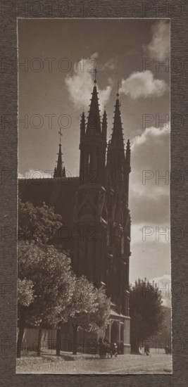 St. Anne's Church; photograph, St. Anne's Church Vilnius, Vilnius Lithuania, 1912; photograph, photographic print: height 16.3 cm, width 7.0 cm; album page: height 31.0 cm, width 23.5 cm, A tall Gothic-style stone building with several pointed spires rises against a sky with scattered clouds; the building appears dark brown and black in the photograph and the sky shows shades of gray and white. In front of the building a group of leafy trees in shades of green and brown line a street area, and a horse-drawn carriage with dark-colored horses and figures is visible near the building’s arched entrance. The overall image is sepia-toned with borders in textured dark brown.