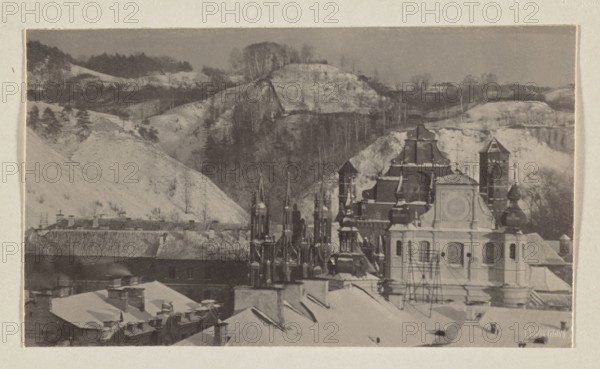 Churches and mountains; photograph from the album Vilnius, volume 1; Jan Bulhak 1876–1950; Vilnius, 1912; scenes: Bernardine Church Vilnius, St. Anne's Church Vilnius, St. Michael's Church Vilnius, panoramas; photographic print: 9.5 × 16.3 cm; album page: 31.0 × 23.5 cm., The image shows a snowy scene with a group of rooftops and ornate church towers in the foreground and a hilly, wooded landscape behind them. Visible colors include shades of gray, off-white, beige, and muted brown. The foreground contains several pitched roofs covered in snow, chimneys, and scaffolding near a light-colored building with decorative architectural details. Clustered behind and beside that building are darker, more ornate spires and domes with layered shapes and pointed tops. In the background a series of pale, snow-covered hills rise, punctuated by darker tree lines and bare trees on the slopes, creating a contrast between the lighter hills and the darker wooded ridges. The whole scene is framed by a pal...
