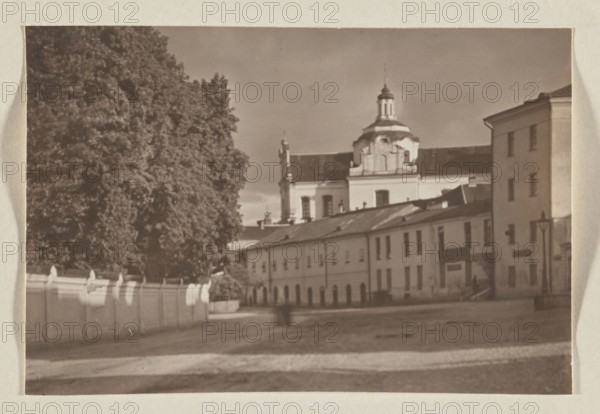 Dominican Church of the Holy Spirit Vilnius, 1912, photograph, photographic print: height 11.2 cm, width 16.6 cm, album page: height 31.0 cm, width 23.5 cm, Vilnius Lithuania, The image shows a sepia-toned outdoor scene with a large group of trees on the left, a pale wall with decorative posts running along the foreground left, and a row of light-colored buildings with pitched roofs occupying the middle and right. A multi-tiered building with a dome and ornamental facade rises behind the row, and a taller plain building stands to the far right. The ground in front appears open and paved, and a few indistinct human shapes are visible near the buildings. The visible colors are shades of brown, beige, and cream.