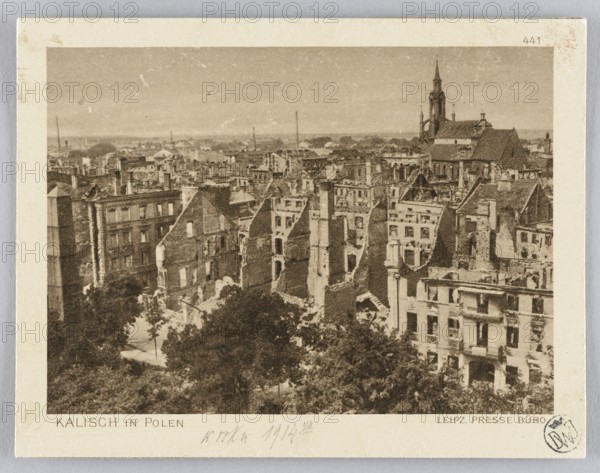 Panorama of Kalisz after its destruction in 1914; panorama city view showing war damage and churches. Leipziger Presse-Büro press office original author; JF Lehmanns Verlag Munich publisher. Postcard glued across the entire surface onto a cardboard backing; postcard, 1914. Germany place of origin; Munich place of publication. Dimensions: height 9.3 cm, width 12.0 cm., A sepia-toned photograph shows a dense urban scene of damaged and partially ruined multi-story buildings with many exposed walls, broken roofs, and missing sections, creating jagged rooflines and interior views; a tall church with a steeple is visible rising above the cluster of buildings in the background; there is a group of trees and foliage in the foreground partially obscuring lower parts of the buildings; the overall palette includes shades of brown, beige, tan, and darker brown tones across the structures and vegetation.