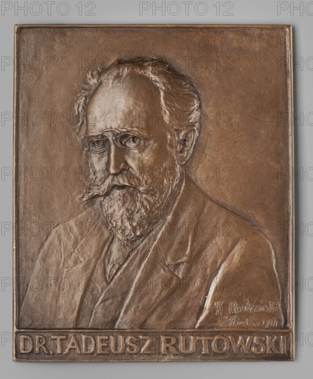 Obv.: male bust turned 3/4 to the left, pince-nez on the nose, slight baldness on the forehead, long moustache, beard with sideburns, jacket on the shoulders; inscription at the bottom in the field: 1916. Portrait plaque of Kazimierz Mieczyslaw Chodzinski 1861–1921 by Dr. Tadeusz Rutowski 1841–1912; male busts; Vienna; dimensions: 126 × 152 mm., A bronze relief portrait of an older man with a beard and mustache wearing round glasses and a suit is shown on a rectangular plaque with a raised border; the background and plaque are various shades of brown and bronze, and the surrounding surface visible around the plaque is gray.