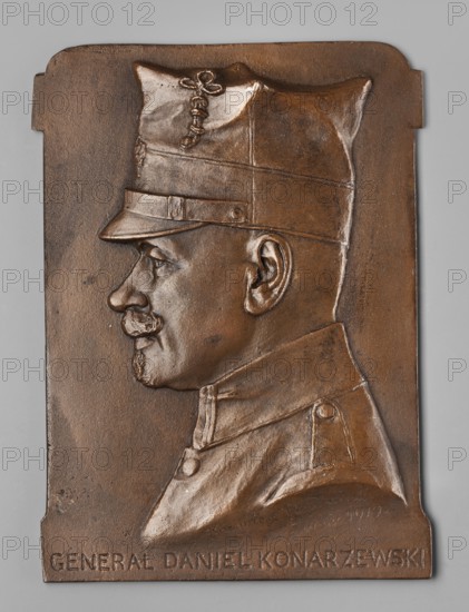 Daniel Konarzewski; Chodzinski, Kazimierz Mieczyslaw 1861–1921 medalist/author; Kupiecki, Jan foundry/co-creator; Chodzinski, Kazimierz 1861–1919. Male busts; iconography by Konarzewski, Daniel 1871–1935. Kupiecki, Jan foundry; Lviv. Obverse: male bust in uniform in left profile, wearing a peaked cap, curled mustache, small beard; inscription at the bottom. 1919, Poland. Plaque. Dimensions: 130 mm; 180 mm., A bronze relief plaque showing the left-profile bust of a man wearing a peaked military-style cap and a high-collared coat; the figure has a mustache and short beard, a visible ear, and defined facial features. The plaque surface is textured and patinated in bronze tones with darker and lighter areas, and it has a rectangular shape with small notches on the sides and rounded corners. The background behind the relief is a smooth neutral gray.