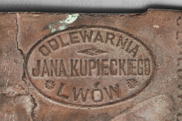 Daniel Konarzewski; Chodzinski, Kazimierz Mieczyslaw 1861–1921 medalist/author; Kupiecki, Jan foundry/co-creator; Chodzinski, Kazimierz 1861–1919. Bronze plaque, 1919. Obverse: male bust in uniform in left profile, wearing a peaked cap, curled mustache and small beard; inscription at bottom. Iconography: male busts. Foundry: Kupiecki, Jan Lviv. Dimensions: 130 mm × 180 mm., A close-up of a weathered oval metal plaque set into a rough surface, showing textured pitting and raised letter shapes; the metal is mainly brown with areas of darker brown and rusty tones, and there are patches of pale green corrosion and small spots of off-white residue. The plaque sits within an indented border and displays worn, embossed shapes and decorative elements, with the surrounding area showing scratches, cracks, and uneven surfaces in similar brown and rust hues. The top edge of the image shows a horizontal strip of darker grey.