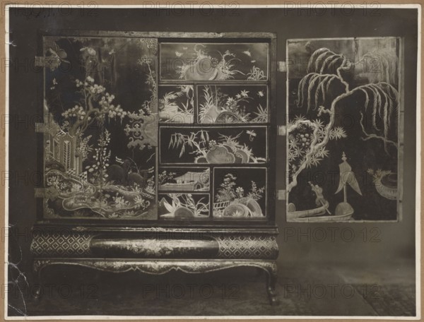 A rectangular two-wing cabinet with drawers inside, circa 1720–1740, French Regency style, chinoiserie, decorated with genre scenes and plant motifs; print glued on toned brown cardboard, ca. 1925 photographic reproduction of the cabinet, photographic print: 16.5 × 21.7 cm; cardboard: 35.0 × 25.1 cm., The image shows a decorative cabinet with ornate panels and an open side door, featuring intricate painted scenes of plants, trees, rocks, and small boats. The cabinet rests on curved legs and a patterned base with floral and geometric designs. Visible are stylized branches, willow-like drooping foliage, and various leafy plants, along with decorative hinges and metal fittings on the doors. The scene decoration includes several compartments each with different landscape motifs and a small figure in a boat on the open panel. Colors visible in the image include shades of black, brown, beige, and cream, with variations of dark and light tones creating contrast across the painted surfaces.