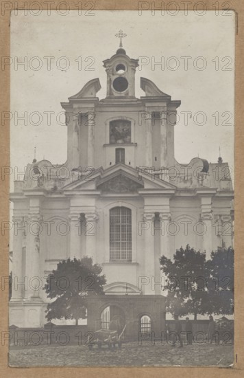 Sienno Sianno, Belarus. Dominican Church of St. Trinity — general view towards the façade, architecture, gates, churches, sacred architecture, lost monuments, general views. Photographer unknown. ca. 1910–1913. Print glued to brown cardboard. Photography from nature; photographic print: 13.8 × 8.7 cm; cardboard: 35.3 × 25.5 cm., A large classical stone building with columns and decorative curved roof elements dominates the image, shown in a muted grayscale. The facade includes a tall central window with a grid of panes, an ornate triangular pediment above it, and a round opening near the top beneath a small tower topped by a cross. There is visible weathering and texture on the building surfaces. In front of the building is a low brick wall with an arched opening flanked by metal gates, and a group of dark-leaved trees on either side. On the cobblestone street in front are a few people standing and walking, and a couple of horse-drawn carts with visible wheels and harnesses. The overal...