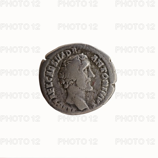 Denarius, Antoninus Pius 86–161; Roman emperor 138–161. Obverse: Head of Antoninus Pius uncovered to the right, inscription IMP T AEL CAES HADRI - ANTONINVS. Reverse: Diana standing to the right, holding a bow in her outstretched left hand and an arrow in her lowered right hand, inscription AVG PIVS PM TR PC - OS DES II. Iconography: standing Diana; attributes: bows, arrows. Date: 138. Place of origin: Rome, Roman Empire. Medium: coin. Diameter: 16.5–18.0 mm. Weight: 2.78 g. Axis: 195°., A circular metallic coin with a worn surface and irregular edge, shown against a plain light background; the coin appears mostly grey with darker grey areas and slight brownish tones, and shows a raised profile of a bearded man's head facing right with visible hair and facial contours, surrounded by a raised border and a ring of indistinct marks around the portrait.