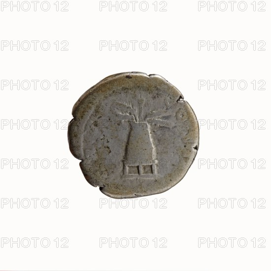 Denarius, Antoninus Pius 86–161; Roman Emperor 138–161. Obverse: Head of Antoninus Pius uncovered facing right, legend ANTONINVS · AVG · PIVS · PP. Reverse: Container modius with ears of corn and poppy-heads, legend TR P · COS · II. Iconography: cereals, ears of corn, modius, poppy-heads. Roman, 139. Rome. Coin; diameter 17.0–18.0 mm, weight 3.37 g, axis 180°., A round, worn coin with an uneven edge showing small nicks and chips, colored mainly silver-gray with areas of darker gray and subtle brownish patina; the coin bears a simple raised design resembling a central vertical form with a flared top and short horizontal base, and faint linear marks radiating from the upper portion; the background of the coin surface is mottled with spots of darker discoloration and slight surface abrasions.