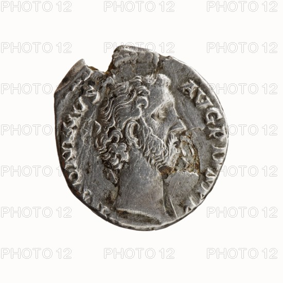 Denarius of Antoninus Pius 86–161; Roman emperor 138–161. Obverse: Head of Antoninus Pius bare, facing right; inscription ANTONINVS · AVG · PIVS · PP. Reverse: Sacred implements from left—knife secespita, sprinkler aspergillum, jug capis, crosier lituus, and ladle simpulum; inscription TR P COS II. Date: 139. Place: Rome, Roman Empire. Medium: silver coin. Diameter: 15.5–17.0 mm. Weight: 3.17 g. Axis: 195°., A circular metallic object with a worn surface showing an embossed profile of a bearded man facing right, with detailed curly hair and facial features; the object has irregular edges and small areas of surface damage and pitting; the visible colors are silver-gray with patches of darker gray and small areas of brownish discoloration.