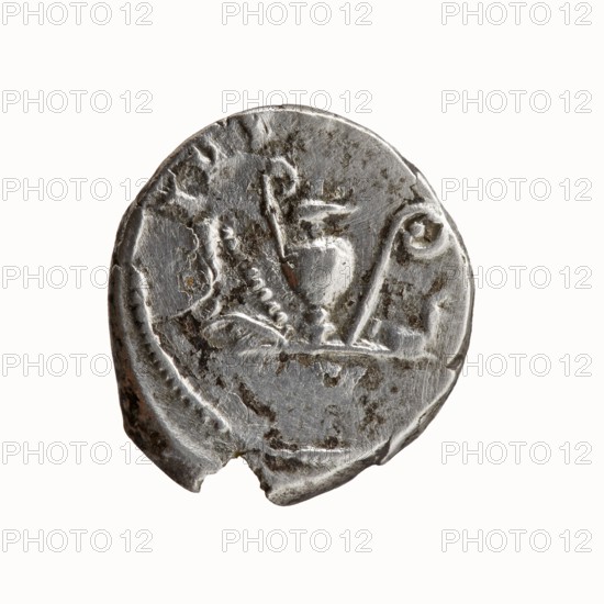 Denarius, Antoninus Pius 86–161; Roman emperor 138–161. Obverse: Head of Antoninus Pius bare, right; legend ANTONINVS • AVG • PIVS • PP. Reverse: Sacred implements from the left — knife secespita, sprinkler aspergillum, jug capis, crosier lituus, and ladle simpulum; legend TR P COS II. Date: 139. Place of origin: Rome, Roman Empire. Medium: coin denarius. Diameter: 15.5–17.0 mm. Weight: 3.17 g. Axis: 195°., A round, worn metal coin with a rough, irregular edge and a small chip on its rim; its surface shows raised, embossed shapes including curved lines and stylized forms that resemble a vessel and hooked elements, with textured areas of wear and small dark patches of tarnish. The visible colors are silver-gray on the raised metal and darker gray to brownish spots in the corroded areas.