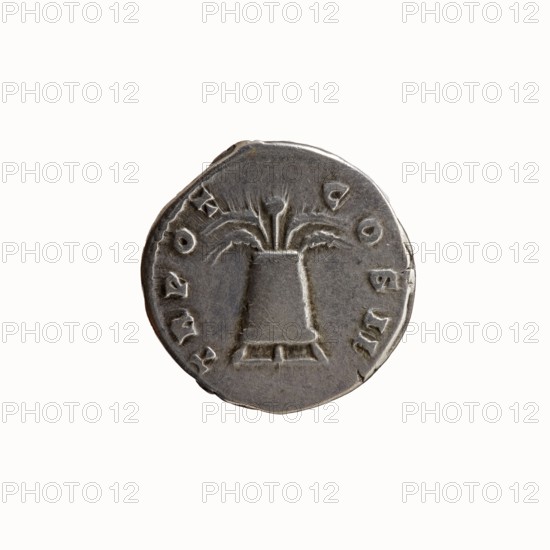 Denarius of Antoninus Pius 86–161; Roman Emperor 138–161. Obverse: Head of Antoninus Pius uncovered facing right, legend ANTONINVS · AVG · PIVS · PP. Reverse: Container modius with ears of corn and poppy head, legend TR POT · COS II. 139 CE. Rome. Silver coin; diameter 17.5 mm, weight 3.58 g, axis 165°., A round metallic object with a worn surface, showing a raised central design resembling a three-pronged floral or fountain-like motif emerging from a conical base, surrounded by shallow indentations and decorative marks along the edge; the object displays shades of gray, silver, and dark gray with subtle highlights and shadows, and the background is off-white.