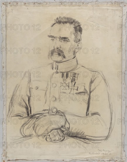 Portrait of Józef Pilsudski, by Józef Mehoffer 1869–1946, half-length drawing of Marshal Józef Pilsudski 1867–1935 with folded hands, made in 1921 in the Belweder Palace corner room., A charcoal or pencil sketch on a pale beige or off-white sheet shows a seated man from the waist up wearing a high-collared uniform with buttons and medals on his chest; he has dark hair and a prominent dark mustache, a composed facial expression, and his arms are folded with his hands resting in front of him. The drawing surface is framed by a rough fabric border with visible frayed edges and stitching in light beige and off-white tones, and the sketching marks vary from soft gray to darker black strokes against the light background.